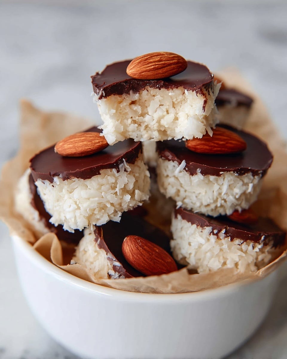 The image shows a small white bowl filled with a pile of round treats. Each treat has two layers: the bottom layer is a textured white coconut base with a rough surface, and the top layer is a smooth, dark brown chocolate disc. On top of each chocolate layer, there is a whole almond placed in the center. The bowl is set on a white marbled surface with a blurred white tile background. photo taken with an iphone --ar 4:5 --v 7