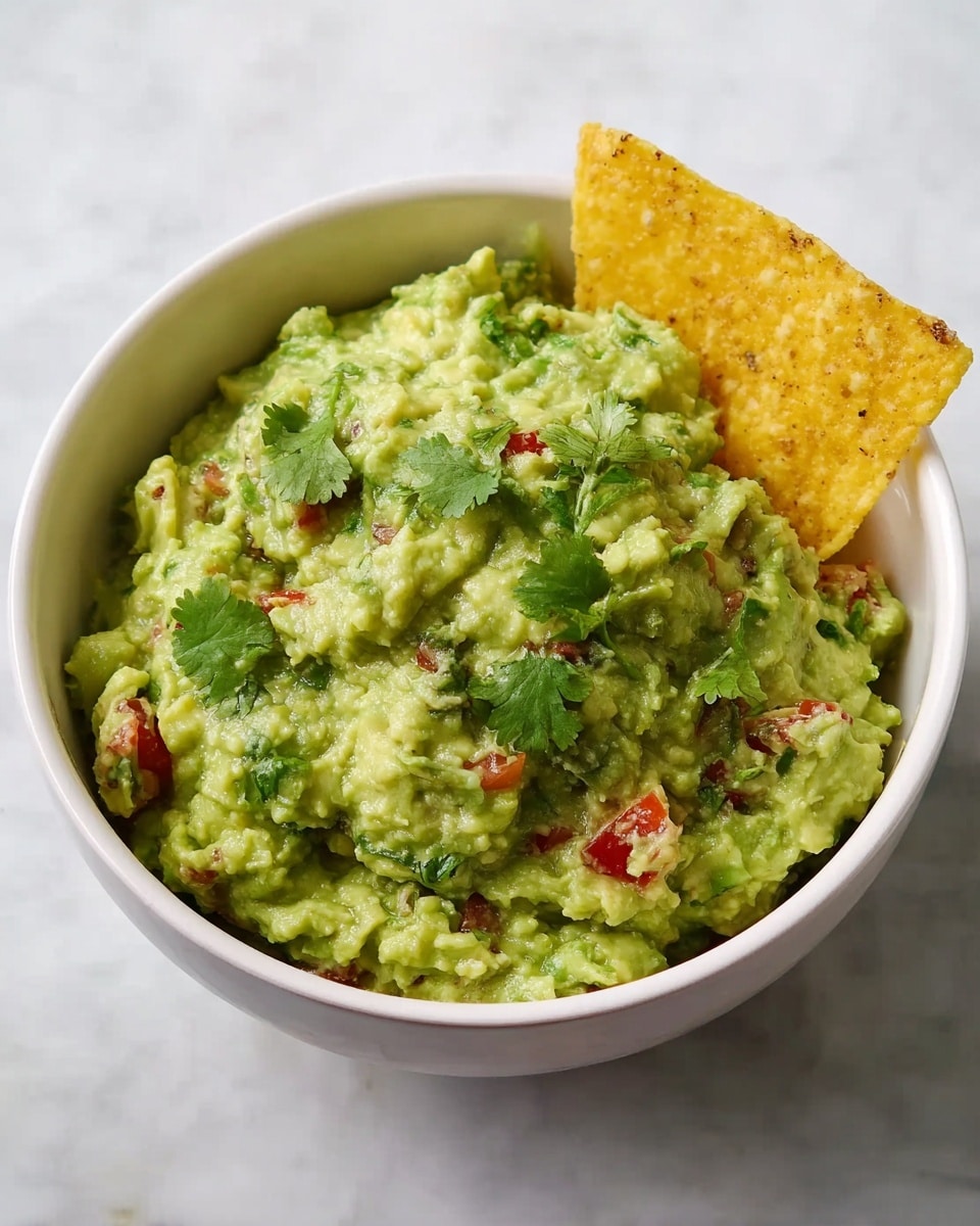 A white bowl filled with chunky green guacamole showing different textures from mashed avocado and small pieces. Mixed inside are small bits of red tomato and green cilantro leaves, with some cilantro sprinkled on top for garnish. A yellow tortilla chip is placed sticking out on the right side of the guacamole. The bowl sits on a white marbled surface. photo taken with an iphone --ar 4:5 --v 7