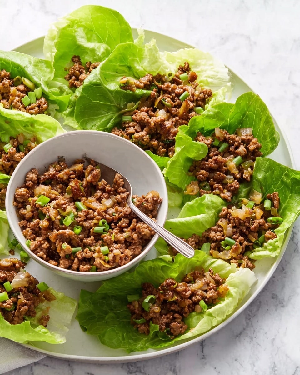 A white round plate with fresh green lettuce leaves arranged around the edges, some filled with a cooked mixture of browned ground meat, light brown diced onions, and small green pieces of scallion on top, positioned on a white marbled surface. In the center of the plate, there is a white bowl filled with the same meat mixture, and a silver spoon is resting in the bowl. The textures show the crispness of the lettuce and the crumbly, soft meat mixture. Photo taken with an iphone --ar 4:5 --v 7