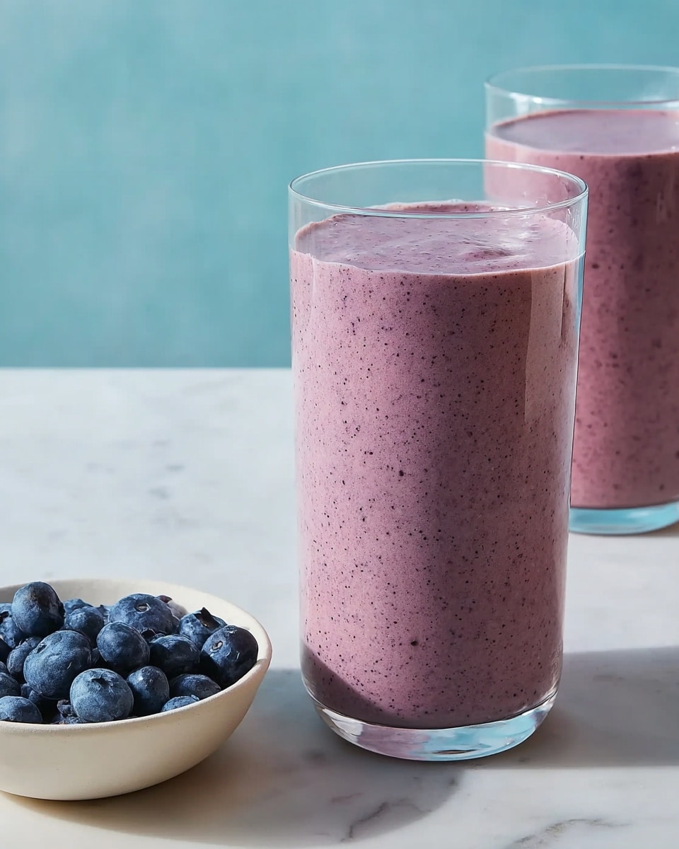 A woman's hand is pouring a thick, creamy pink smoothie with visible small dark specks from a clear ribbed blender container into a clear glass. The smoothie has a smooth but slightly textured surface, filling the glass evenly. Both the glass and blender are placed on a white marbled surface, with a clean, minimal background. Photo taken with an iphone --ar 4:5 --v 7