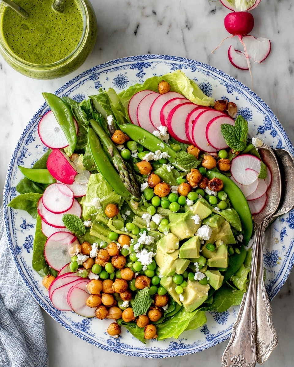A fresh salad is shown in a white bowl with blue flower patterns, filled with several layers of bright green leafy lettuce and mint leaves as the base. On top, there are sliced pink and white radishes, small red radishes, and chunks of light green avocado. Green peas and asparagus pieces add more texture, while golden brown roasted chickpeas add a crunchy contrast. Small bits of white cheese are scattered evenly over the salad. A vintage silver spoon rests on the right side inside the bowl, all placed on a white marbled surface. photo taken with an iphone --ar 4:5 --v 7