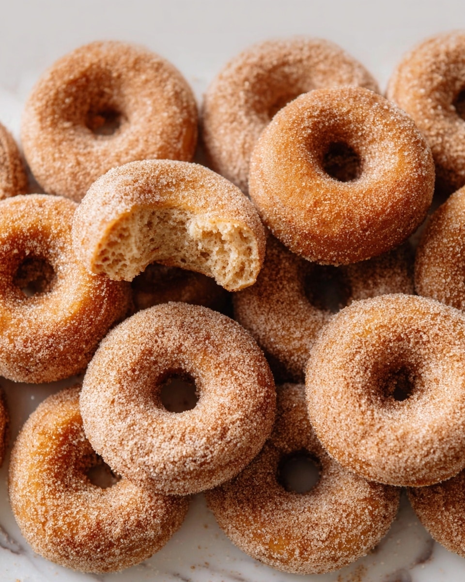 The image shows a close-up of several cinnamon sugar donuts stacked closely together on a white marbled surface. Each donut has a light brown color with a grainy texture of cinnamon sugar coating all around. One donut in the center is broken in half, revealing a soft, slightly crumbly inside with a similar brown shade. The donuts have a classic ring shape with a hole in the middle, and the sugar coating creates a slightly frosted look on the surface. photo taken with an iphone --ar 4:5 --v 7