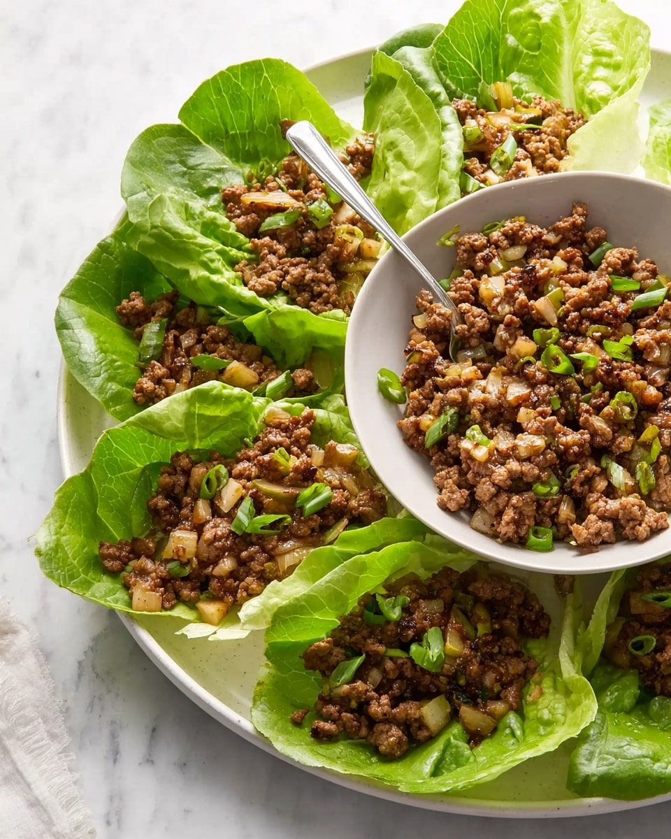 This image shows a white round plate with a white bowl in the center filled with cooked ground meat mixed with small pieces of green vegetables and light-colored onions. The bowl is surrounded by large green lettuce leaves, and some of the meat mixture is placed on these leaves, creating small lettuce cups. The textures show a mix of soft cooked meat and crunchy fresh vegetables. A silver fork rests inside the bowl, and the whole scene is set on a white marbled surface. Photo taken with an iphone --ar 4:5 --v 7
