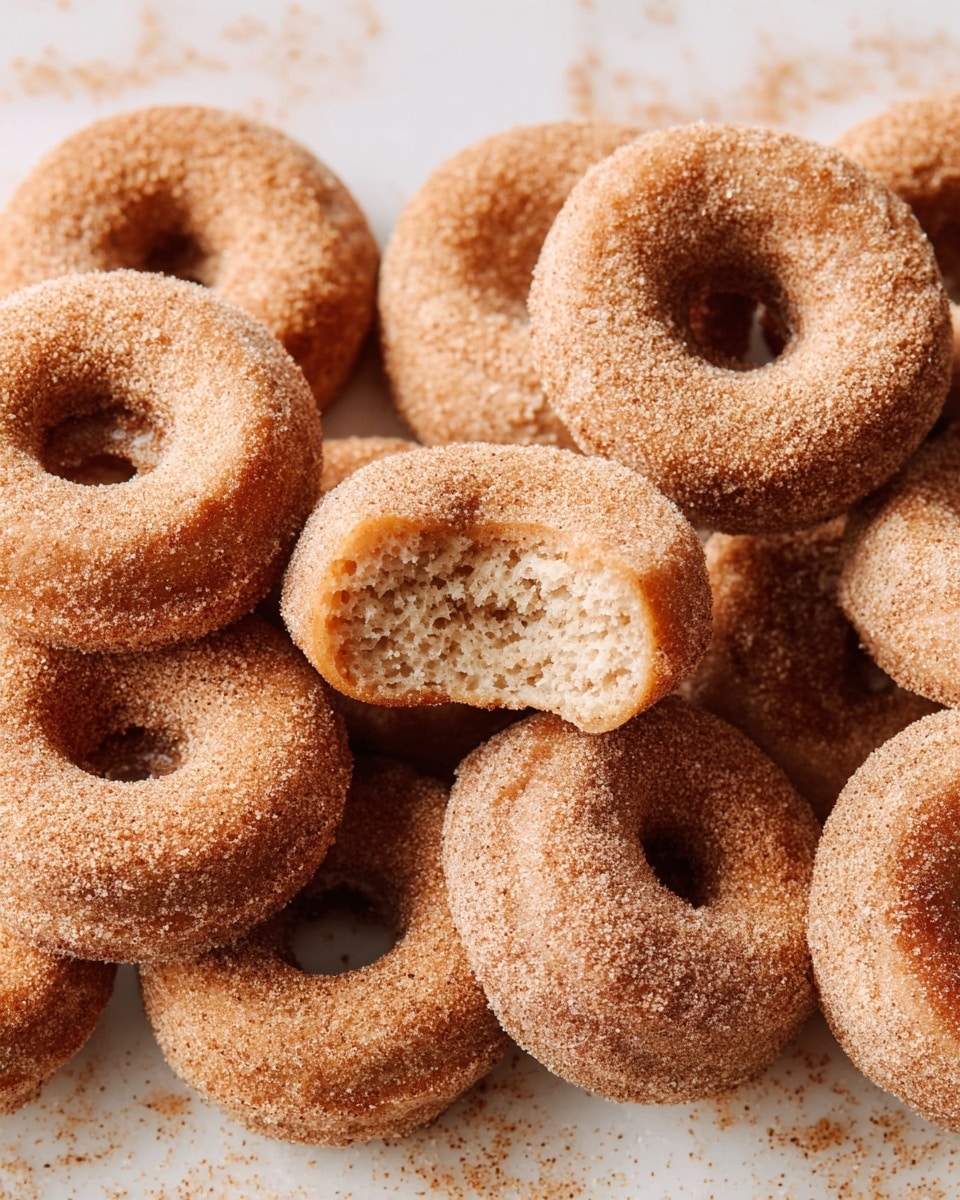 A close-up view of several small cinnamon sugar donuts stacked closely together on a white marbled surface, each donut having a light brown color with a generous layer of grainy cinnamon sugar coating giving them a slightly rough texture; one donut in the center is split in half showing a soft, spongy inside with a light beige interior and sugar crystals on the outside layers; the overall arrangement shows donuts overlapping each other with visible round holes in the middle, giving a warm and inviting look. photo taken with an iphone --ar 4:5 --v 7