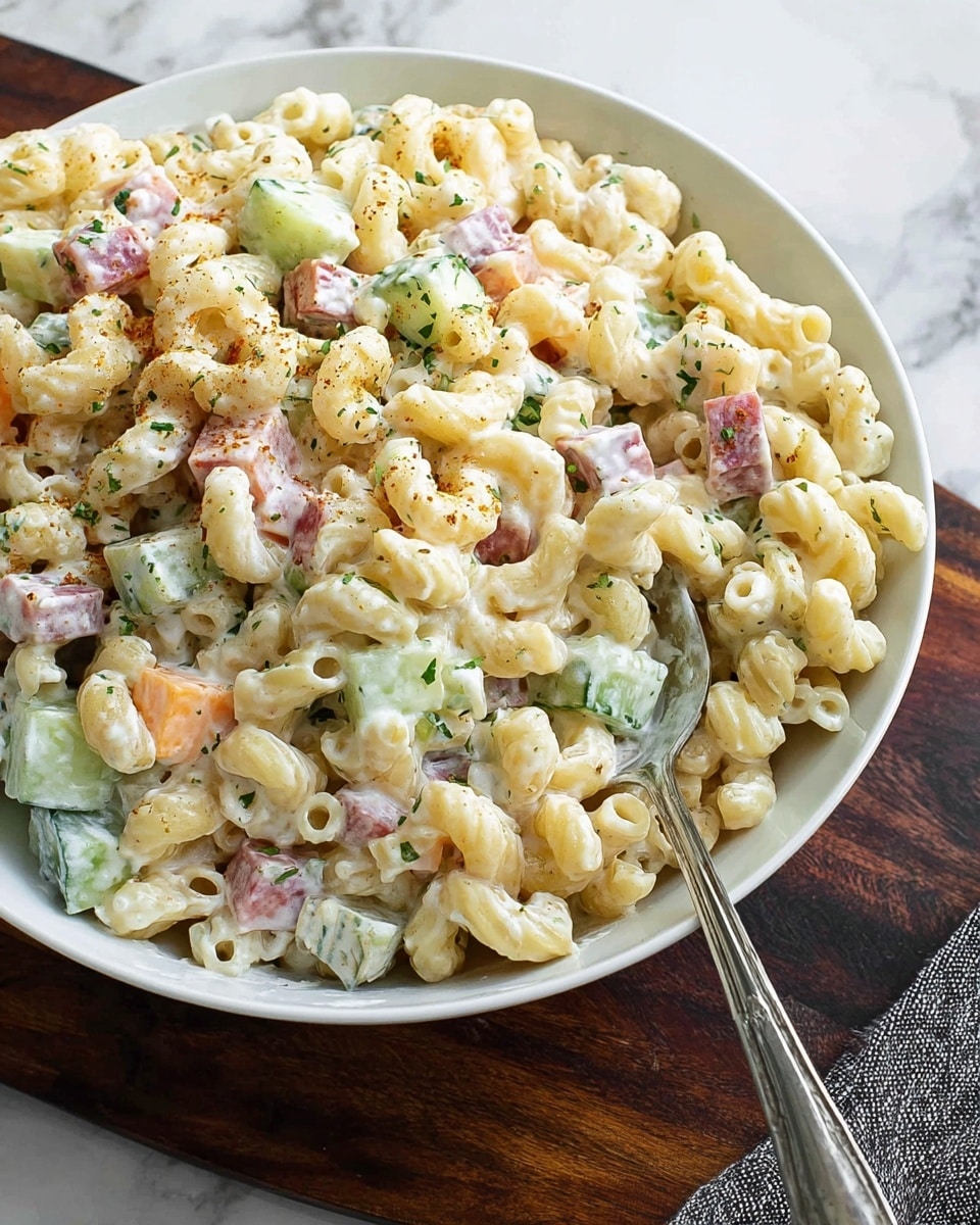 A white bowl filled with creamy macaroni salad showing three main layers: the pale yellow curved pasta forming the base, small cubes of light orange cheese and pink chunks of ham mixed evenly throughout the pasta, and thin green cucumber pieces scattered inside. The dish is coated in a white creamy dressing with green herb specks evenly spread across. A vintage silver spoon sits inside the bowl, resting on the left side. The bowl is placed on a white marbled surface with a white cloth partially visible on the left and some salt crystals near the top edge. Photo taken with an iphone --ar 4:5 --v 7