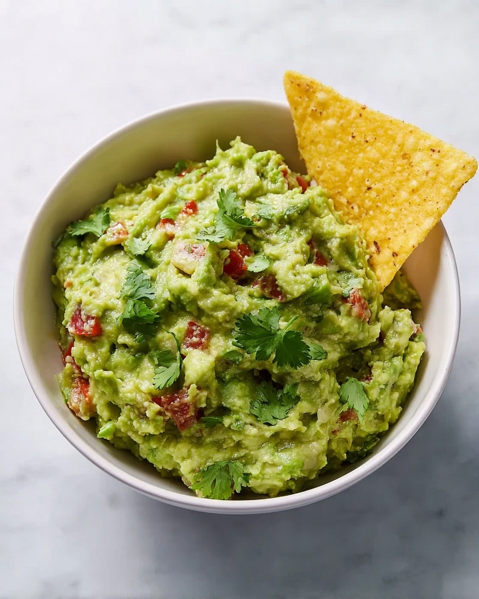 A white bowl filled with green guacamole that has a chunky texture mixed with small red tomato pieces and green cilantro leaves scattered on top. A single yellow tortilla chip is placed upright on the right side of the guacamole. The bowl sits on a white marbled surface. photo taken with an iphone --ar 4:5 --v 7