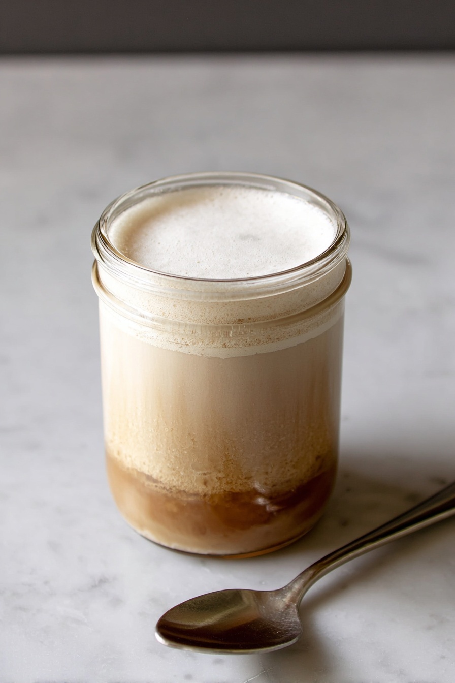 Iced High Protein Coffee, healthy iced coffee, protein coffee recipe, energizing coffee drinks, quick protein coffee - A clear glass jar filled with a two-layer drink sitting on a white marbled surface. The bottom layer is a light brown liquid with some uneven darker spots near the base, making the texture look a bit thick and rich. Above it, a thick, creamy off-white layer fills the top half of the jar, with a smooth frothy surface covering the drink. To the right side of the jar lies a shiny metal spoon, reflecting light. The background and surface are white marble, giving a clean and simple look. Photo taken with an iphone --ar 2:3 --v 7