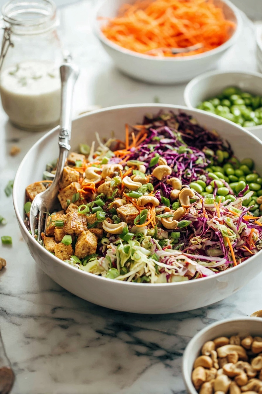 Cashew Crunch Salad with Sesame Dressing, healthy crunchy salads, easy vegetable salads, flavorful sesame salad dressing, vegetarian salad recipes - A large white bowl filled with a colorful salad made of multiple layers including shredded purple and white cabbage, green edamame beans, orange shredded carrots, small chunks of cooked light brown tofu, chopped green onions, and scattered cashew nuts. The salad shows bright, fresh colors with a mix of crunchy and soft textures. A metal fork is placed inside the bowl. In the background, there are two more white bowls, one filled with more shredded carrots and the other with cashew nuts, placed on a white marbled surface. A glass jar with white dressing is also visible. Photo taken with an iphone --ar 2:3 --v 7