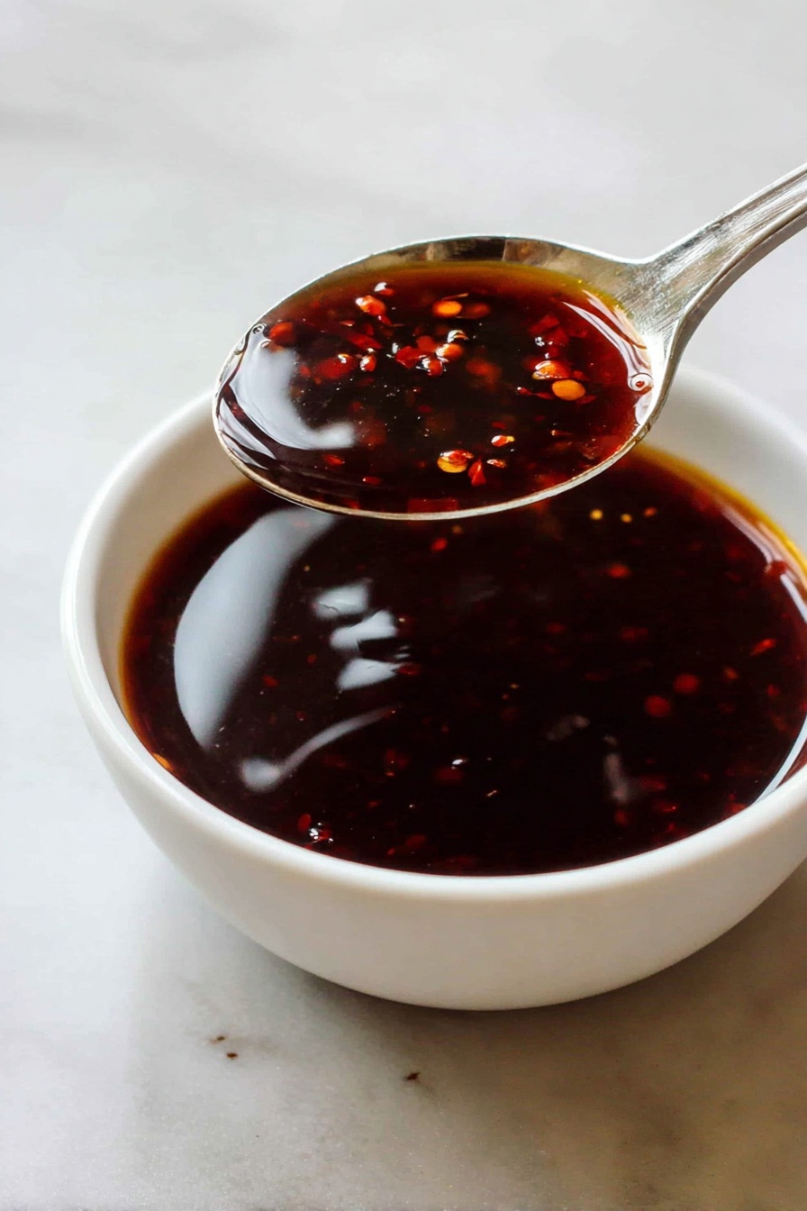 Asian Zing Sauce for Chicken Wings, spicy wing sauce, quick wing sauce recipe, Asian-inspired wing glaze, sweet and spicy chicken wing sauce - A close-up of a white bowl filled with a dark reddish-brown sauce that has a glossy, smooth texture with small bits floating inside. A silver spoon is held above the bowl by a woman's hand, showing the thick, shiny sauce dripping slightly from the spoon's surface. The background is a white marbled texture. photo taken with an iphone --ar 2:3 --v 7