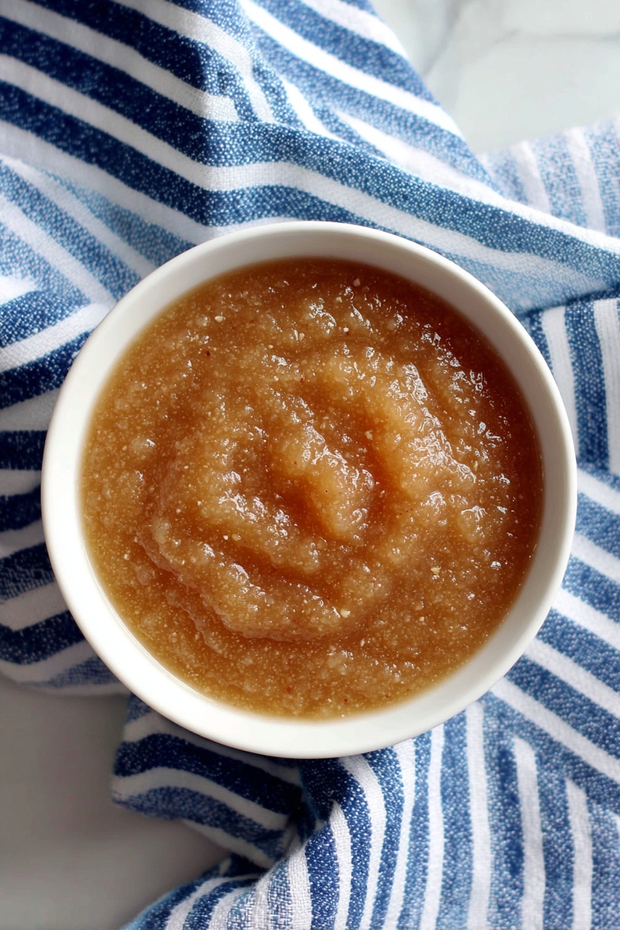 Instant Pot Applesauce, homemade applesauce recipe, easy applesauce recipe, healthy applesauce, quick apple sauce - A white bowl filled with a thick, light brown apple sauce showing small bits and a slightly shiny, wet texture, placed on a blue and white striped cloth. The bowl sits on a white marbled surface, with a whole green apple in the background to the left and a silver spoon partially visible on the right side. The apple sauce looks soft and smooth with some chunkiness visible. Photo taken with an iphone --ar 2:3 --v 7